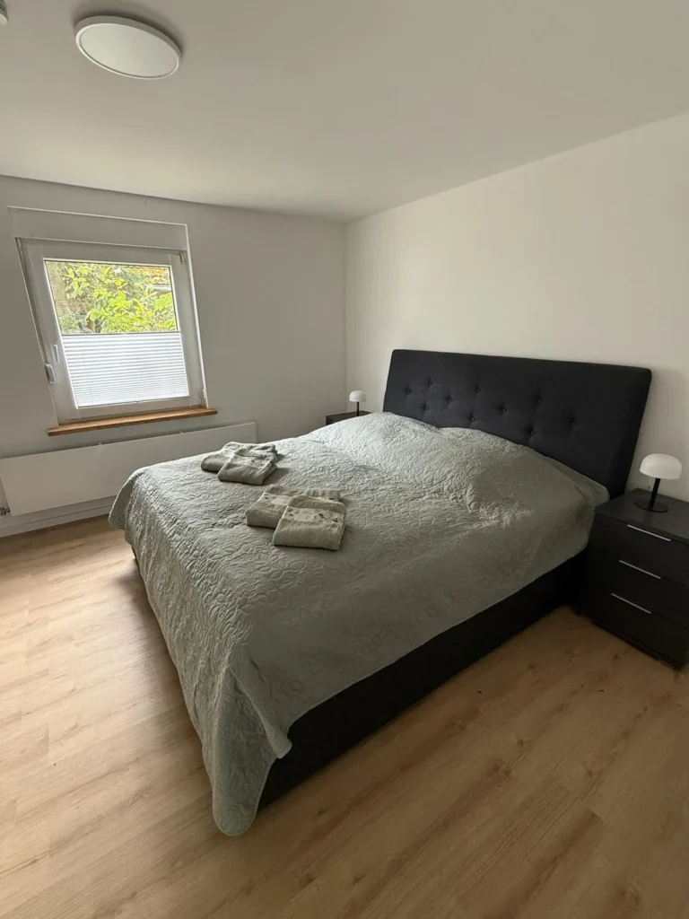 Schlafzimmer im Erdgeschoss der Ferienwohnung mit Doppelbett, gepolstertem Kopfteil, Nachttischen und Fenster mit Plissee
